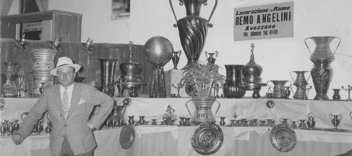 Foto storica: Remo Angelini accanto ai propri lavori in rame esposti, con l'insegna storica "Lavorazione in Rame Remo Angelini"
